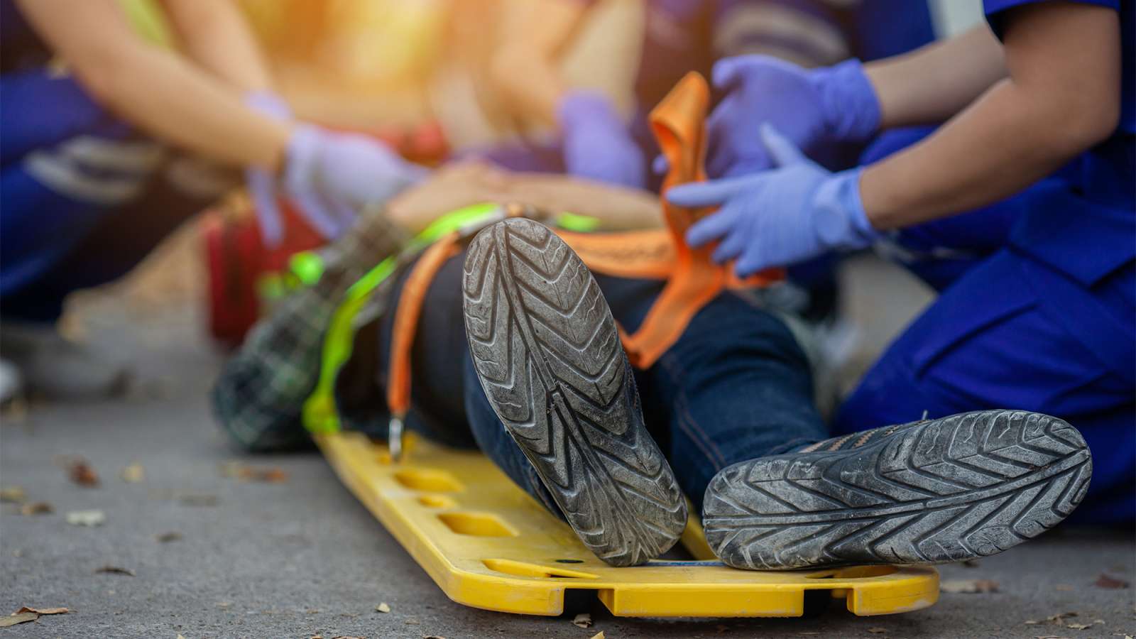 Injured person lying on a yellow spine board on pavement, black-soled sneakers visible, being helped by paramedics.