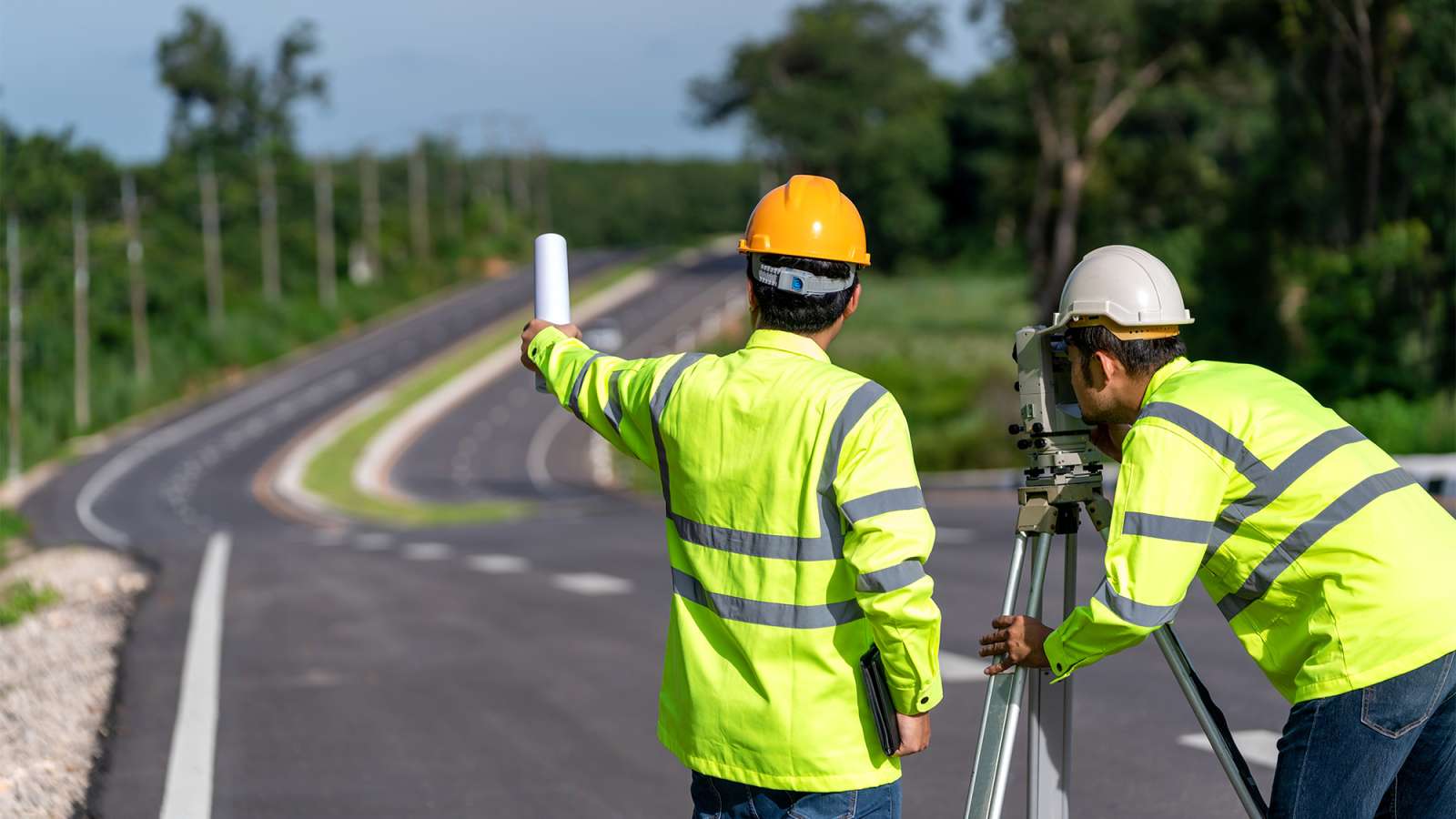 Two road surveyors in hi-vis jackets and hard hats, one using a tripod theodolite, the other gesturing toward a paved road.