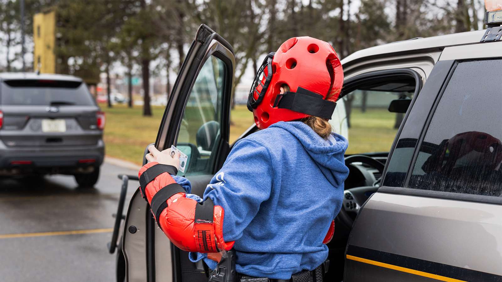 Person wearing red helmet and matching elbow pads over a blue jacket opening a dark SUV door in a tree-lined parking lot.
