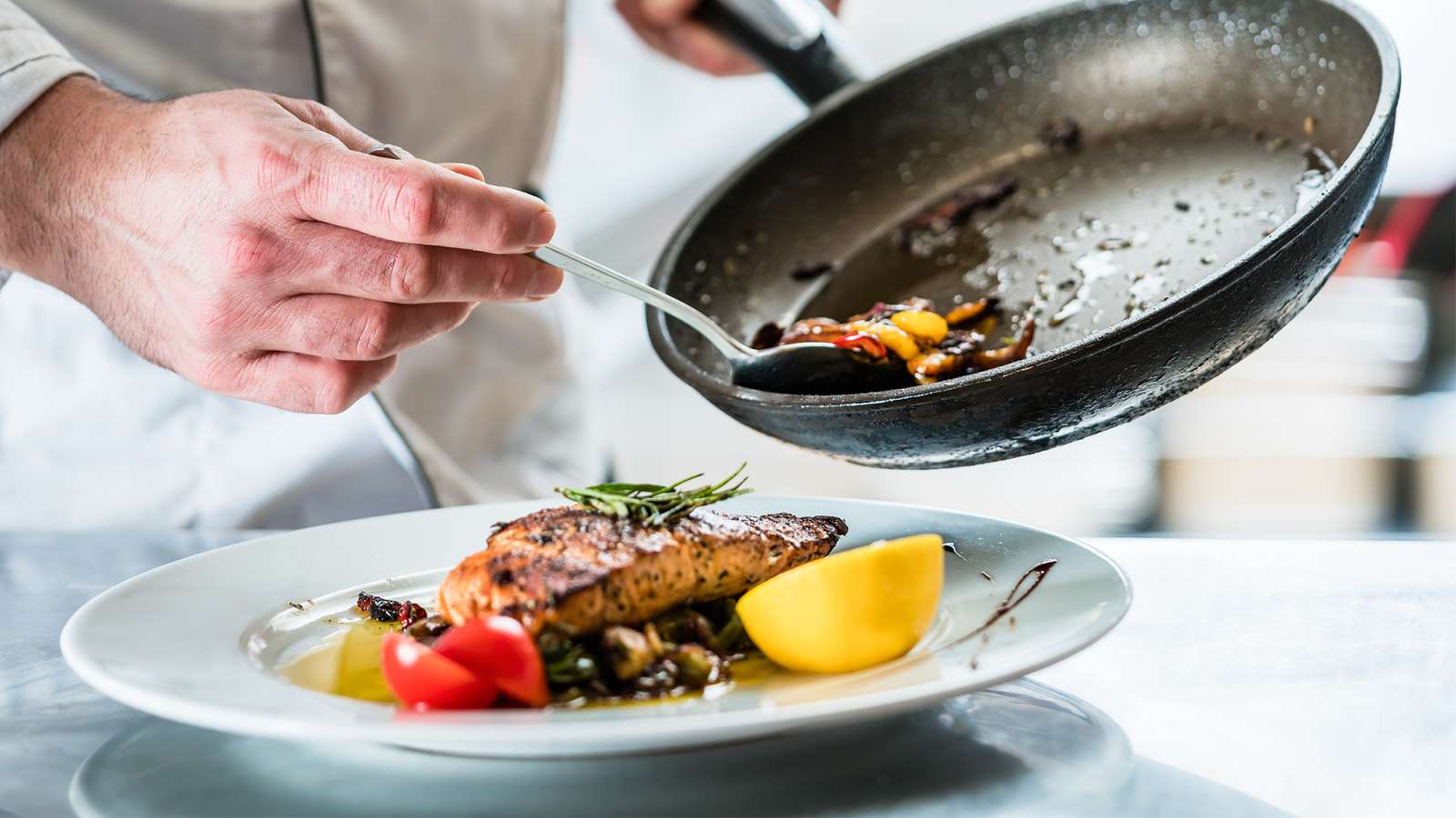 Chef's hands pouring sauce from frying pan onto seared steak with lemon wedge, cherry tomatoes and greens on a white plate.