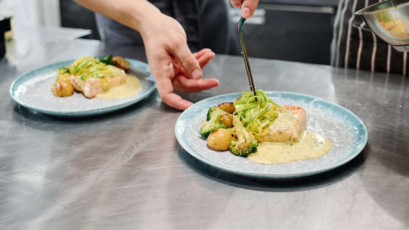 Chef's hands plating seared fish fillets with roasted potatoes, asparagus and creamy sauce on blue ceramic plates.