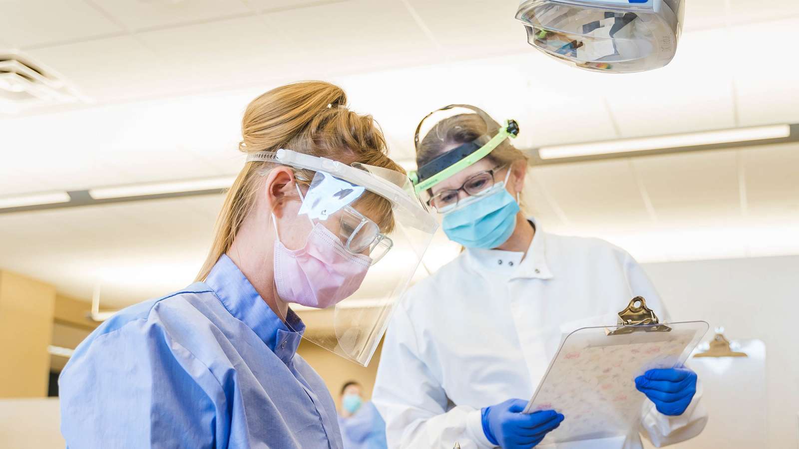 Two masked healthcare workers in face shields and blue gowns reviewing a clipboard in a bright clinical room.