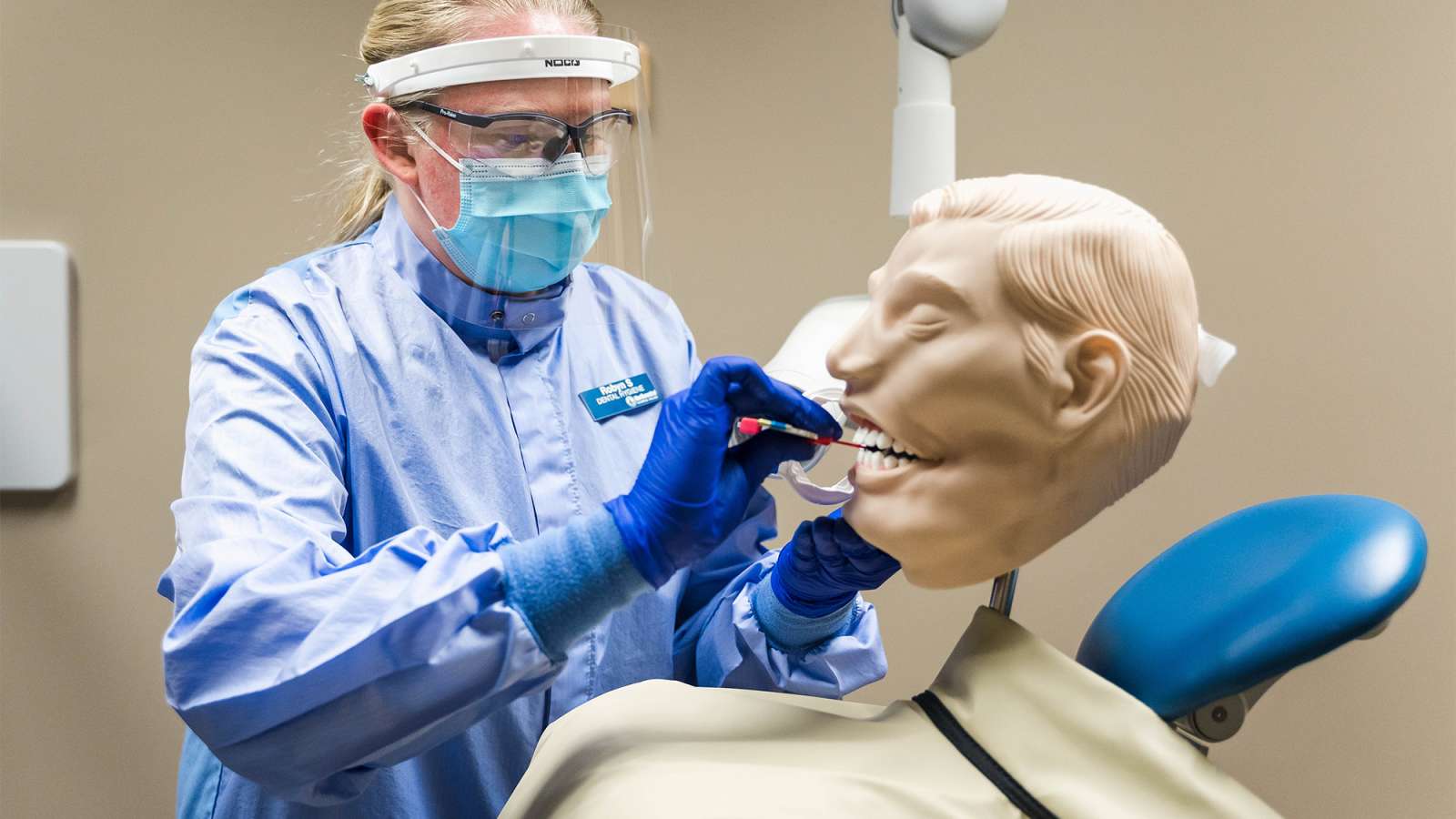 Healthcare worker in blue gown, face shield and mask performing dental procedure on oversized mannequin head.