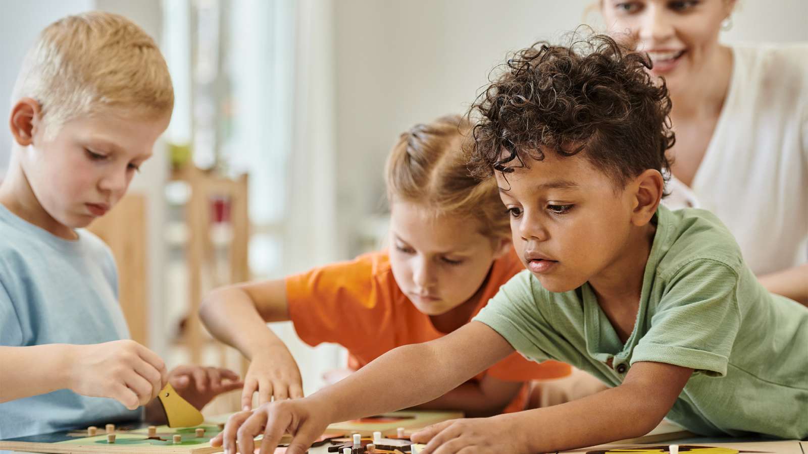 Three young children leaning over a table, intently assembling colorful craft pieces while a smiling teacher watches.