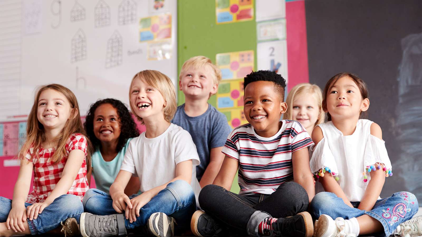 Seven smiling elementary-age children of diverse backgrounds sit cross-legged on a classroom floor in front of a chalkboard.