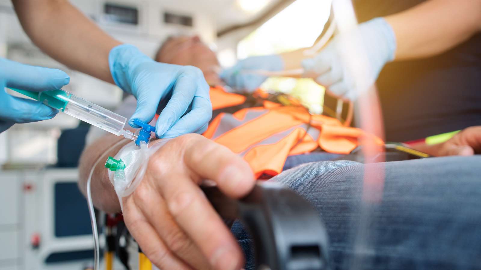 Gloved paramedics administer an IV into a patient's hand on a stretcher inside an ambulance.