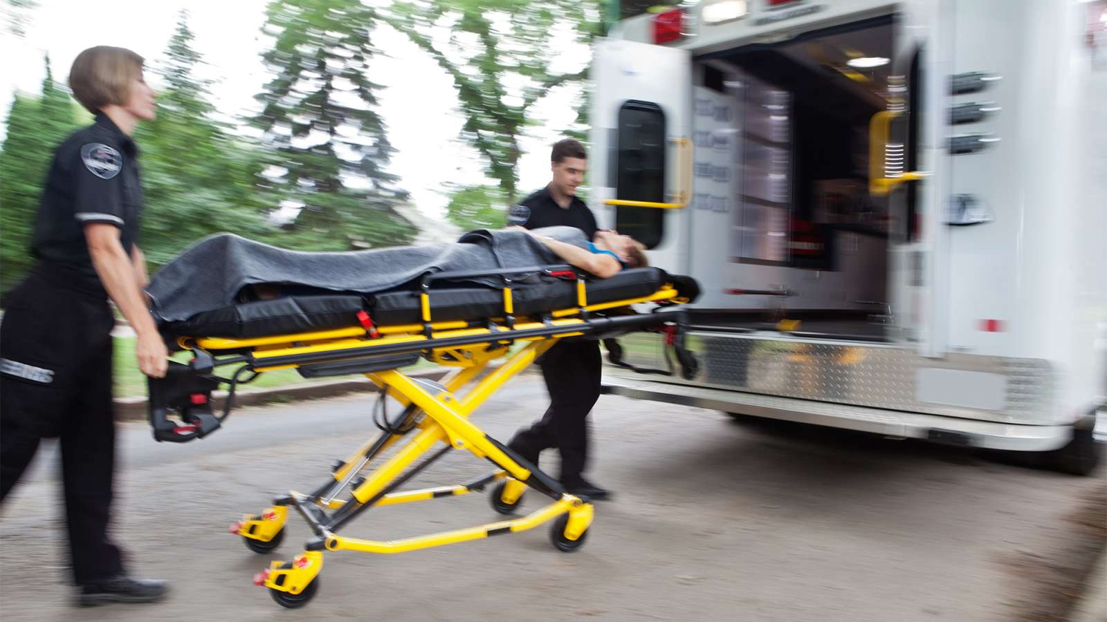 Two uniformed paramedics wheeling a yellow stretcher carrying a person toward the open rear of a white ambulance outdoors.