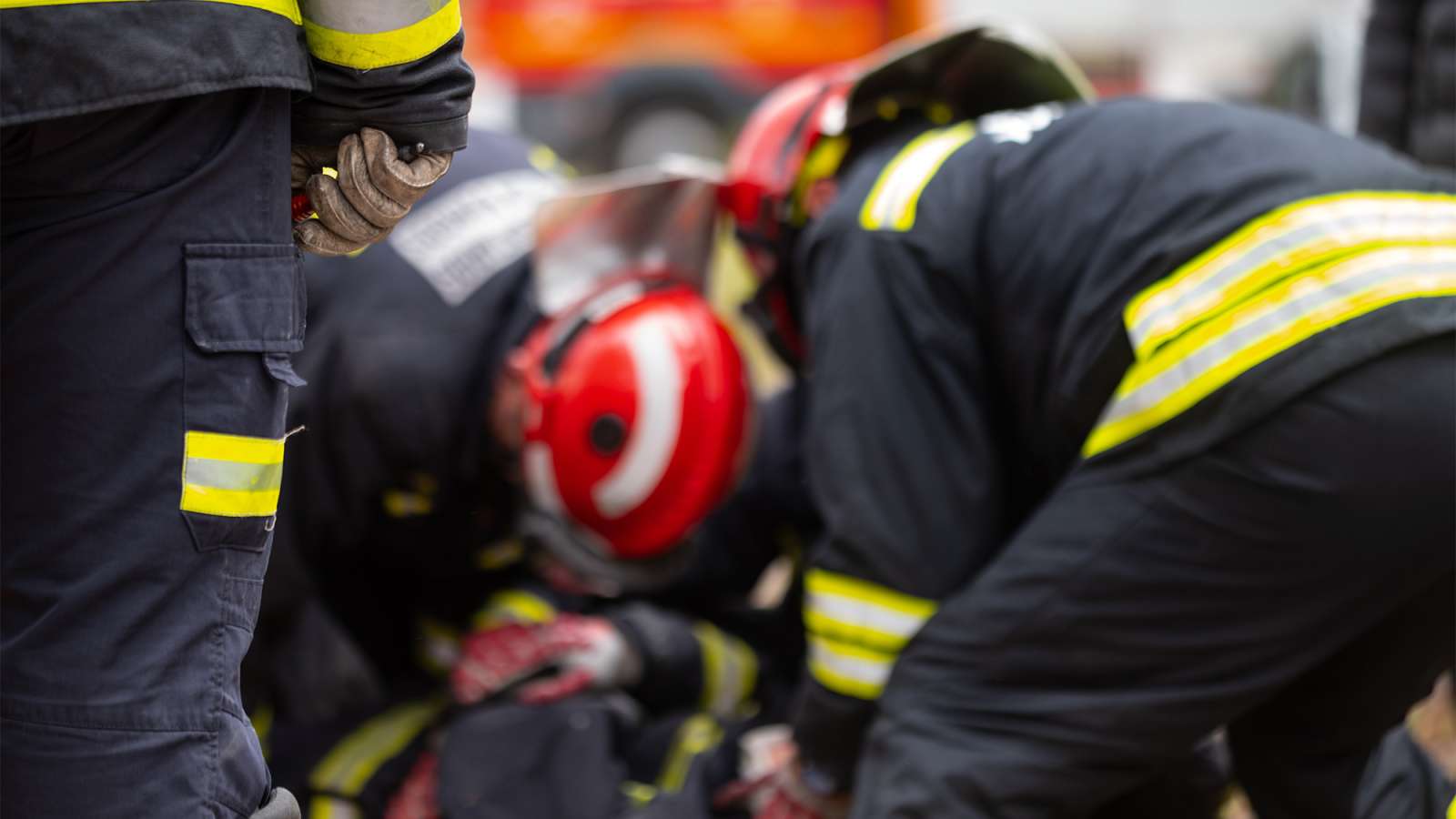 Firefighters in black turnout gear with reflective yellow stripes and a red helmet kneel attending a casualty on the ground.