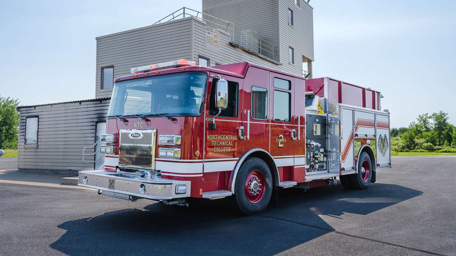 Red fire engine with chrome bumper, ladder and hose compartments parked in front of a modern fire station.