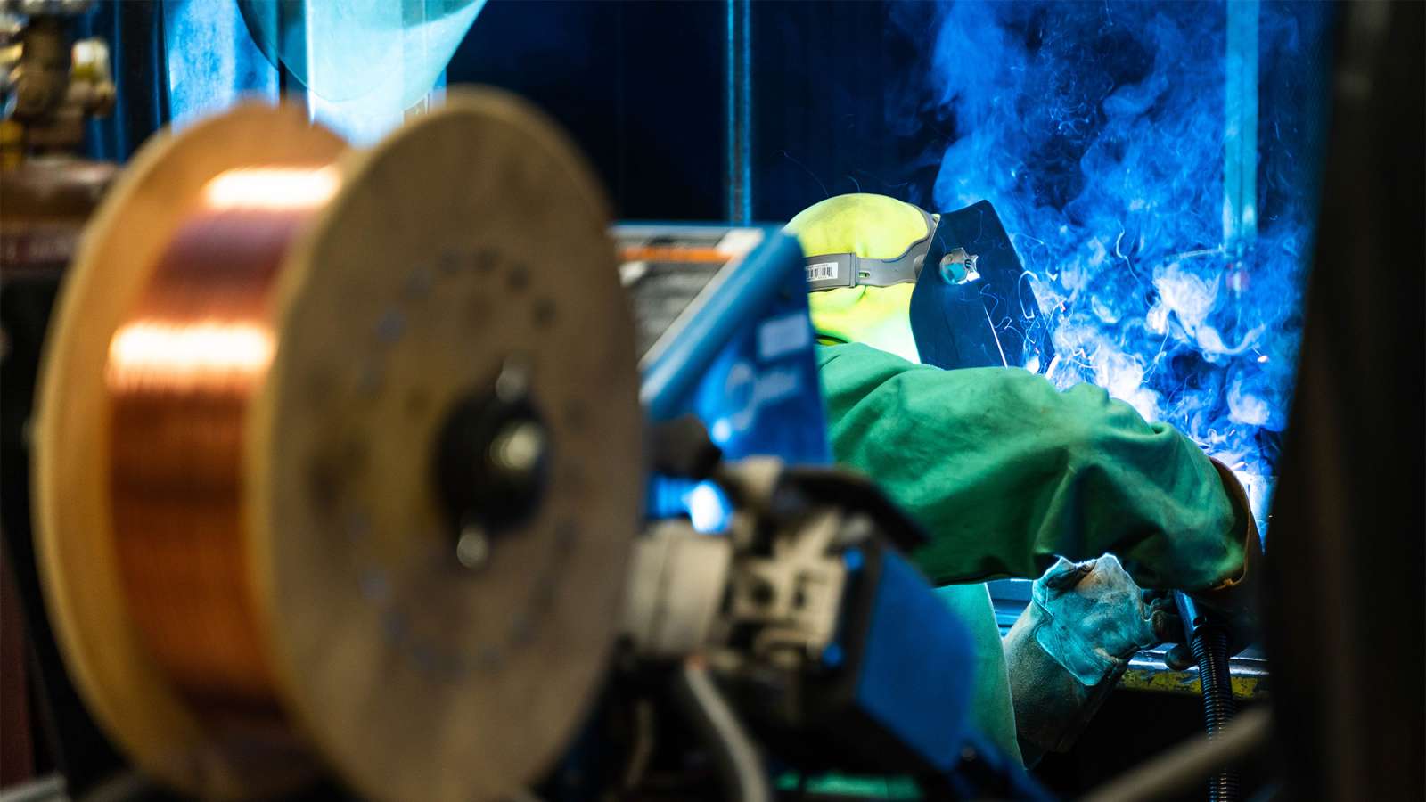 Welder in green protective gear and yellow helmet bent over a joint with a bright blue welding arc and smoke.