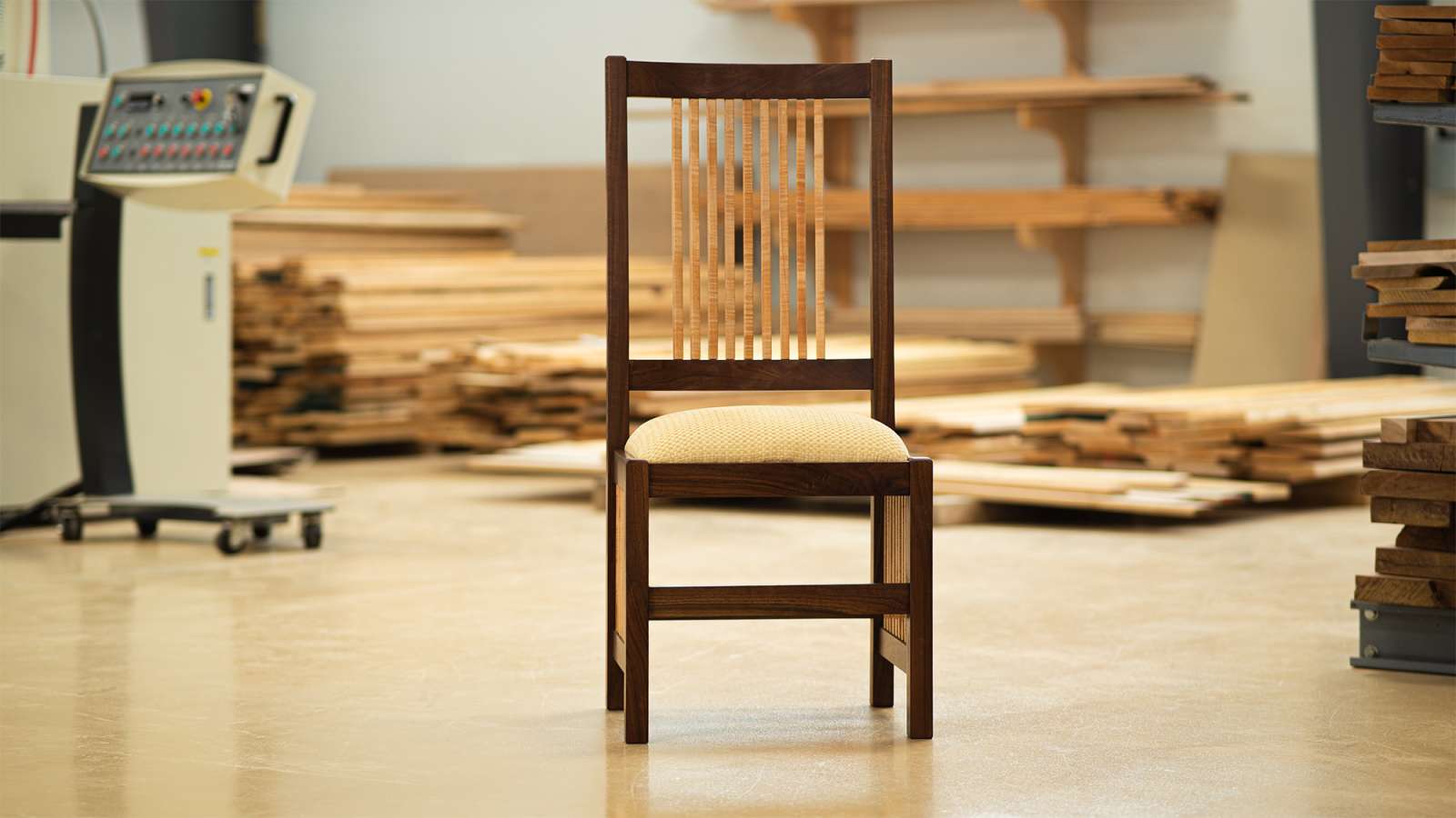 Simple wooden chair with slatted back centered on a workshop floor, surrounded by stacked lumber, and a stationary saw.