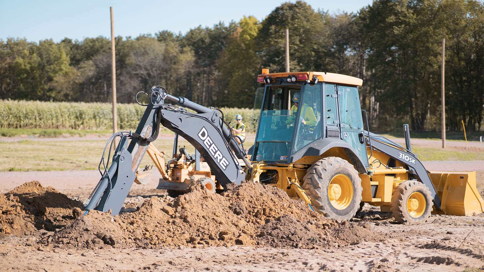 Yellow backhoe loader with extended digging arm scooping a mound of dirt at a rural construction site.