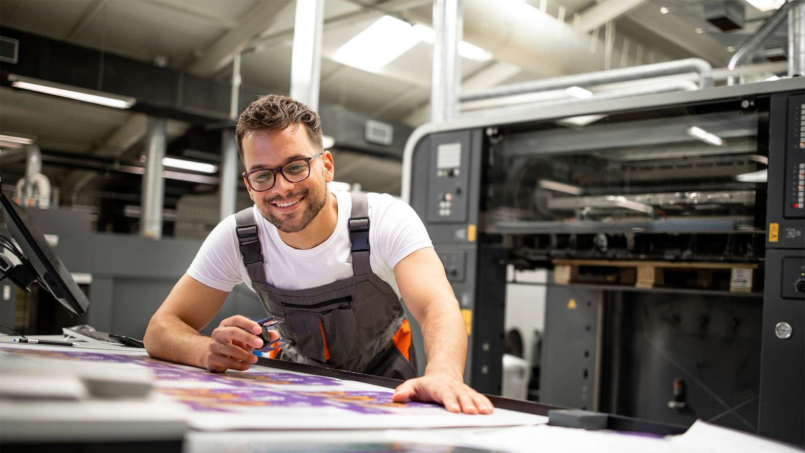 Smiling man with glasses and apron leans over a table of colorful printed sheets beside an industrial printing press.