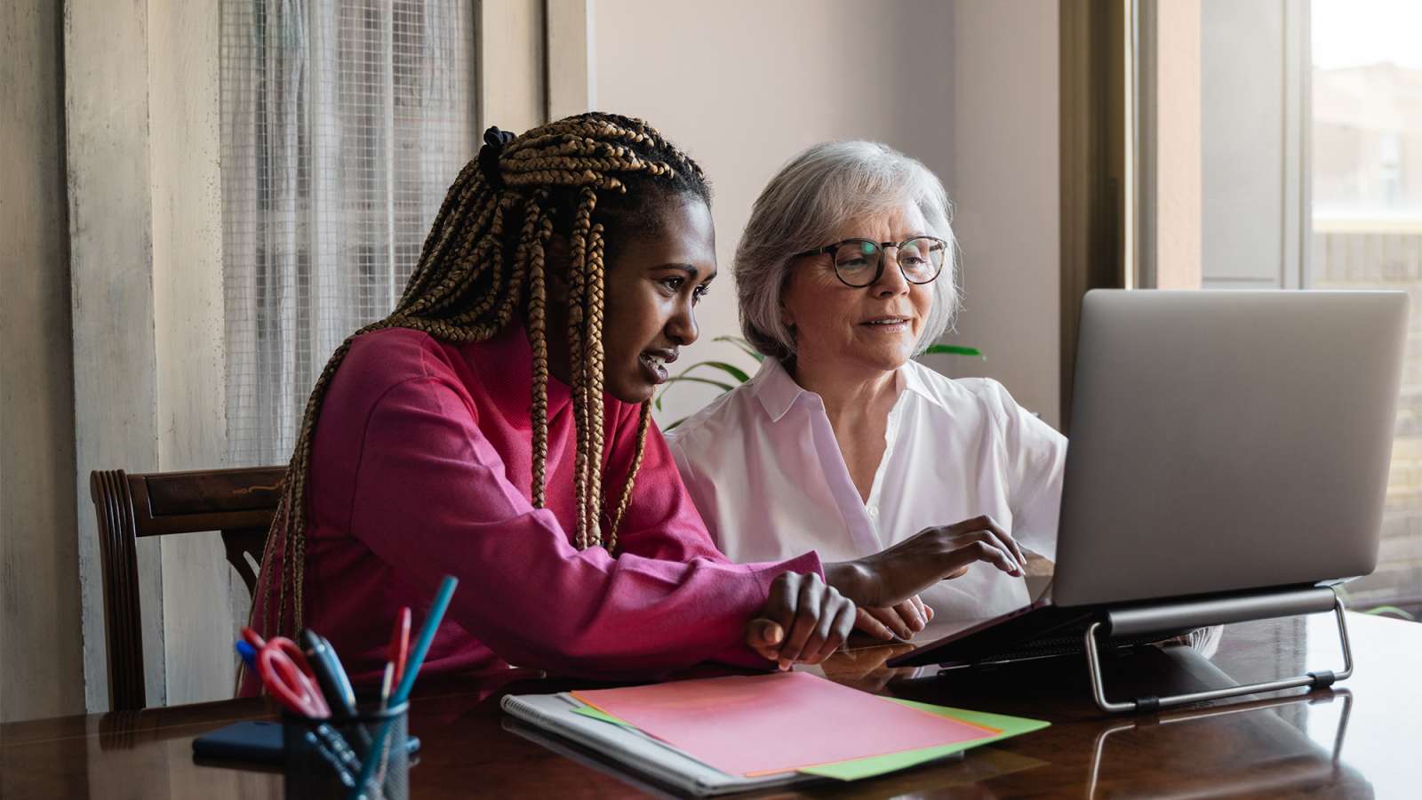 Younger woman in pink sweater points at laptop while an older gray-haired woman with glasses, sitting at a sunlit desk.