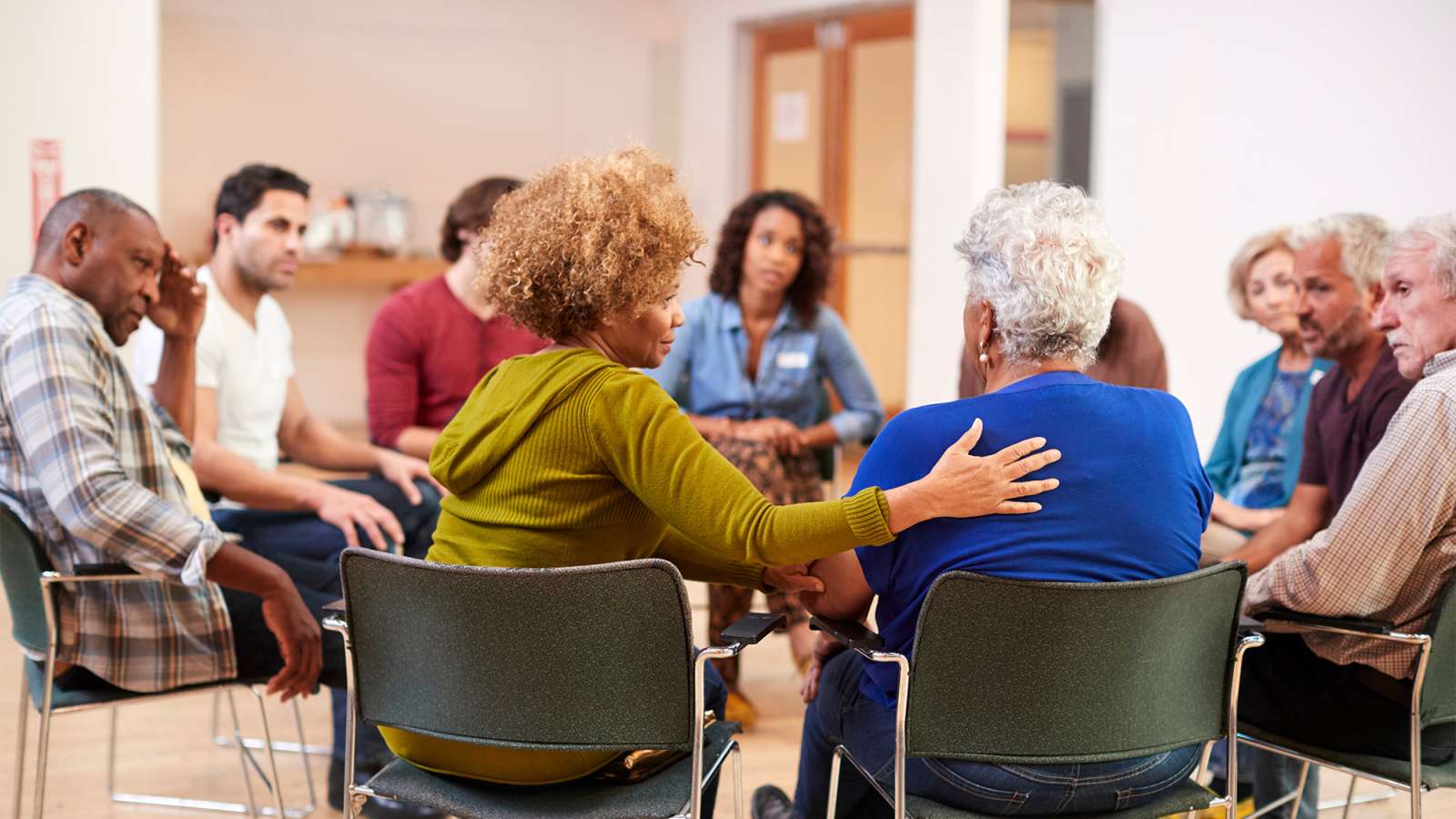 Diverse group of adults seated in a circle in a bright community room; one facilitator comforts an older woman.