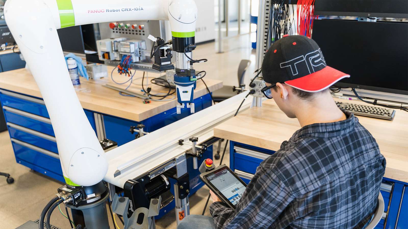 Person in plaid shirt and backward baseball cap seated at a lab bench programming a white robotic arm.