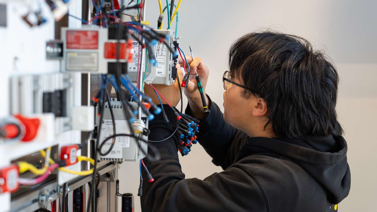 Technician in dark hoodie and glasses carefully connects colorful wires on an electronics test panel in a bright lab.
