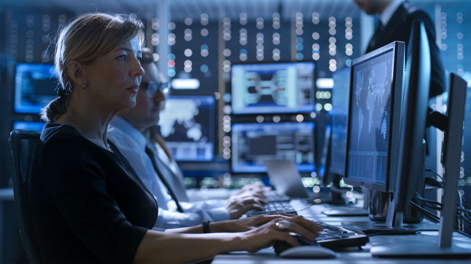 Woman in foreground typing at a multi-monitor security operations desk, focused; colleague blurred in background.