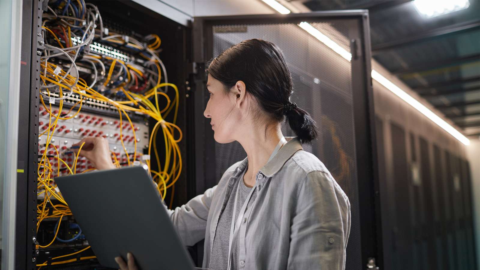 Female IT technician holding a laptop, inspecting organized yellow network cables inside a dimly lit server room.