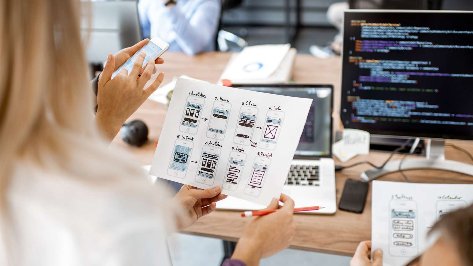 Team gathers around a desk, hands holding printed wireframe sketches and a smartphone while a laptop displays code.
