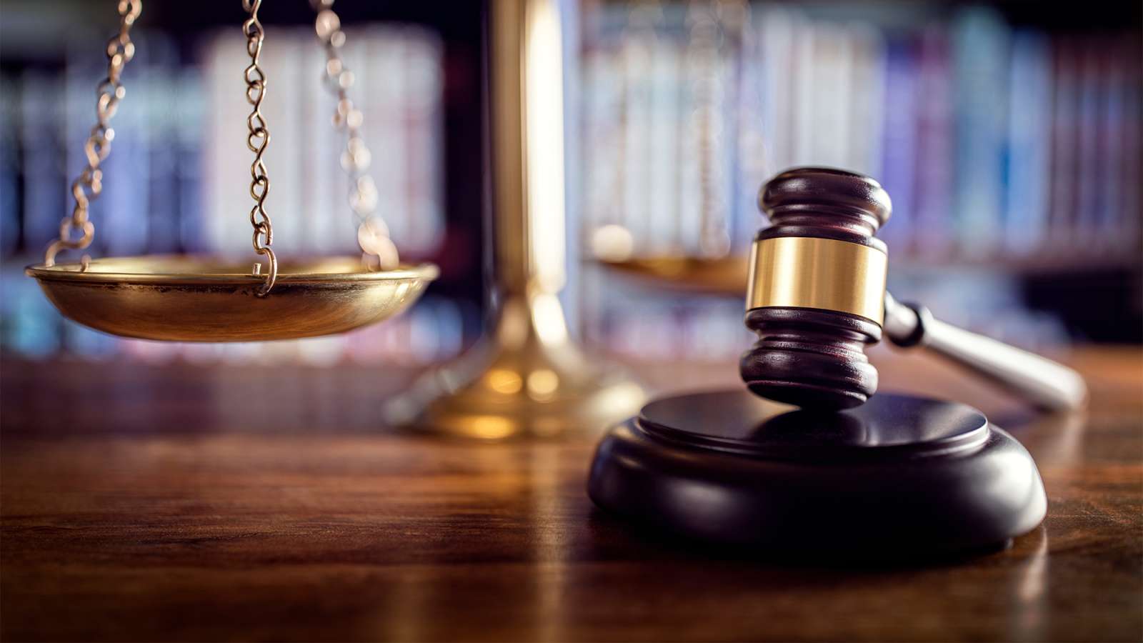 Wooden judge's gavel resting on block beside brass scales of justice, blurred law books on shelf in warm courtroom lighting.