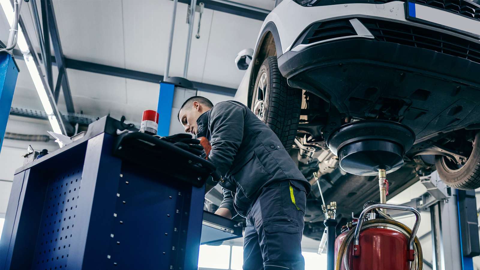 Mechanic in dark coveralls using a laptop at a workstation beside a car raised on a hydraulic lift.