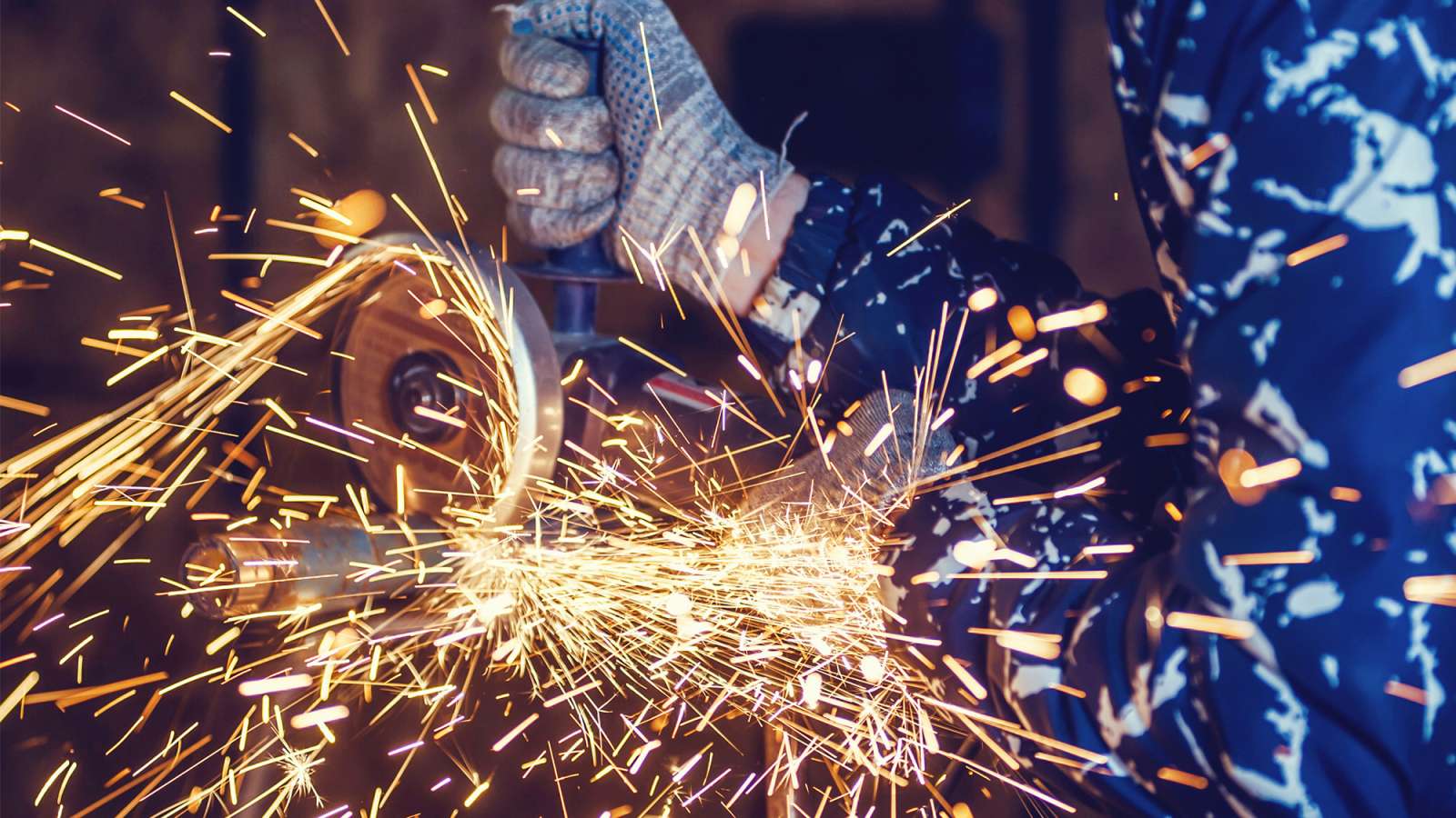 Gloved hands using an angle grinder on metal, showering bright orange sparks against dark blue protective clothing.