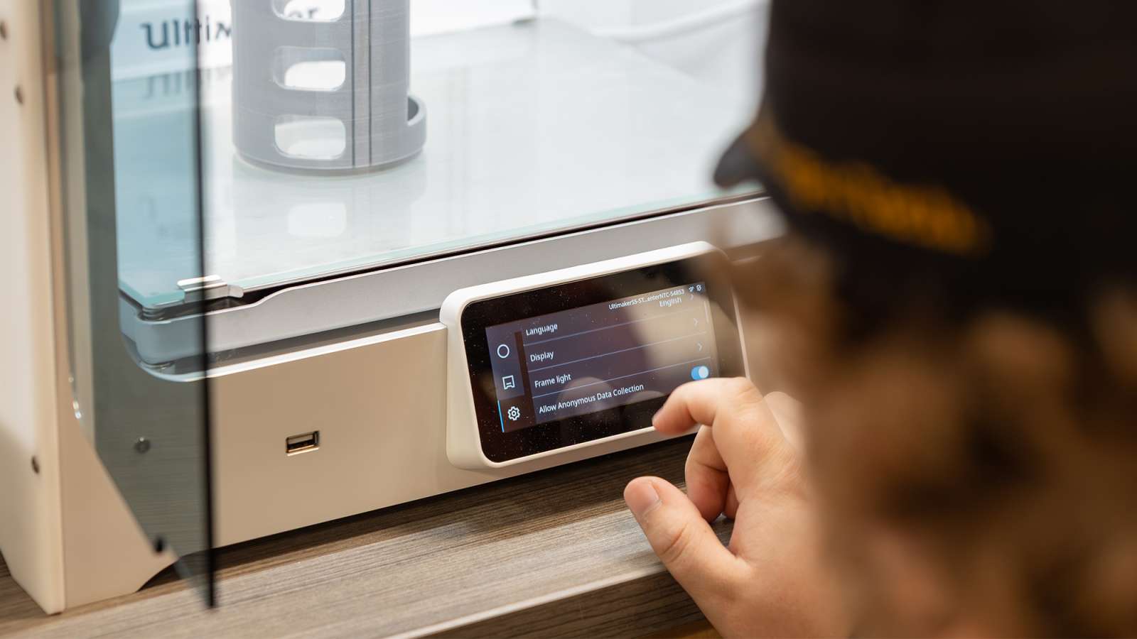 Person’s hand and blurred face interacting with a touchscreen on a desktop 3D printer inside a clear enclosure.