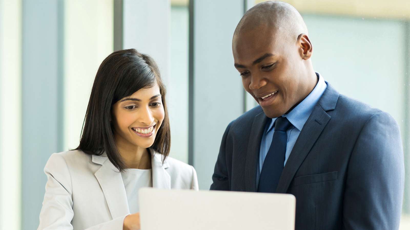 Smiling female and male colleagues in business attire reviewing a laptop together in a bright modern office.