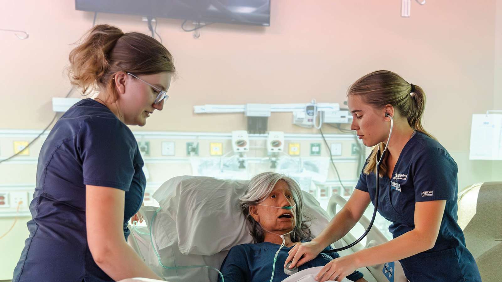 Elderly female patient reclined in hospital bed receiving attentive care from two nurses in navy scrubs—one.