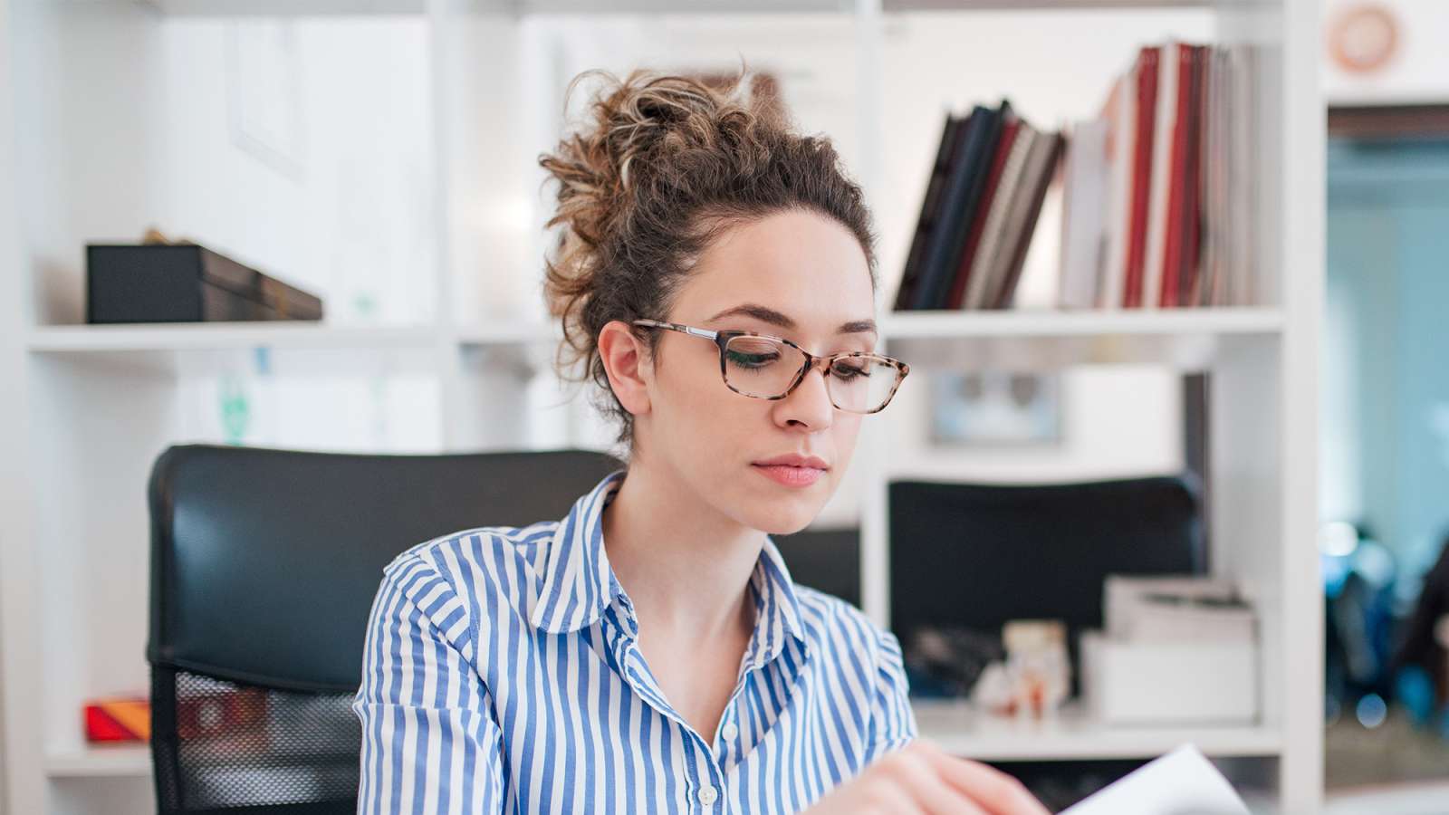 Young woman with curly hair in a bun and glasses, wearing a blue striped blouse, seated at a desk in a bright office.