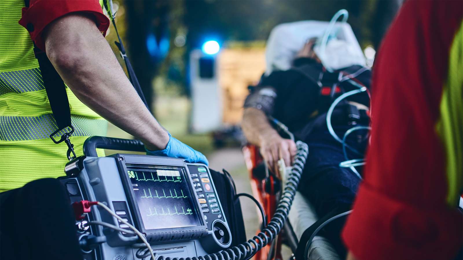 Paramedics wearing hi‑vis vests operate a portable cardiac monitor/defibrillator beside a stretcher.
