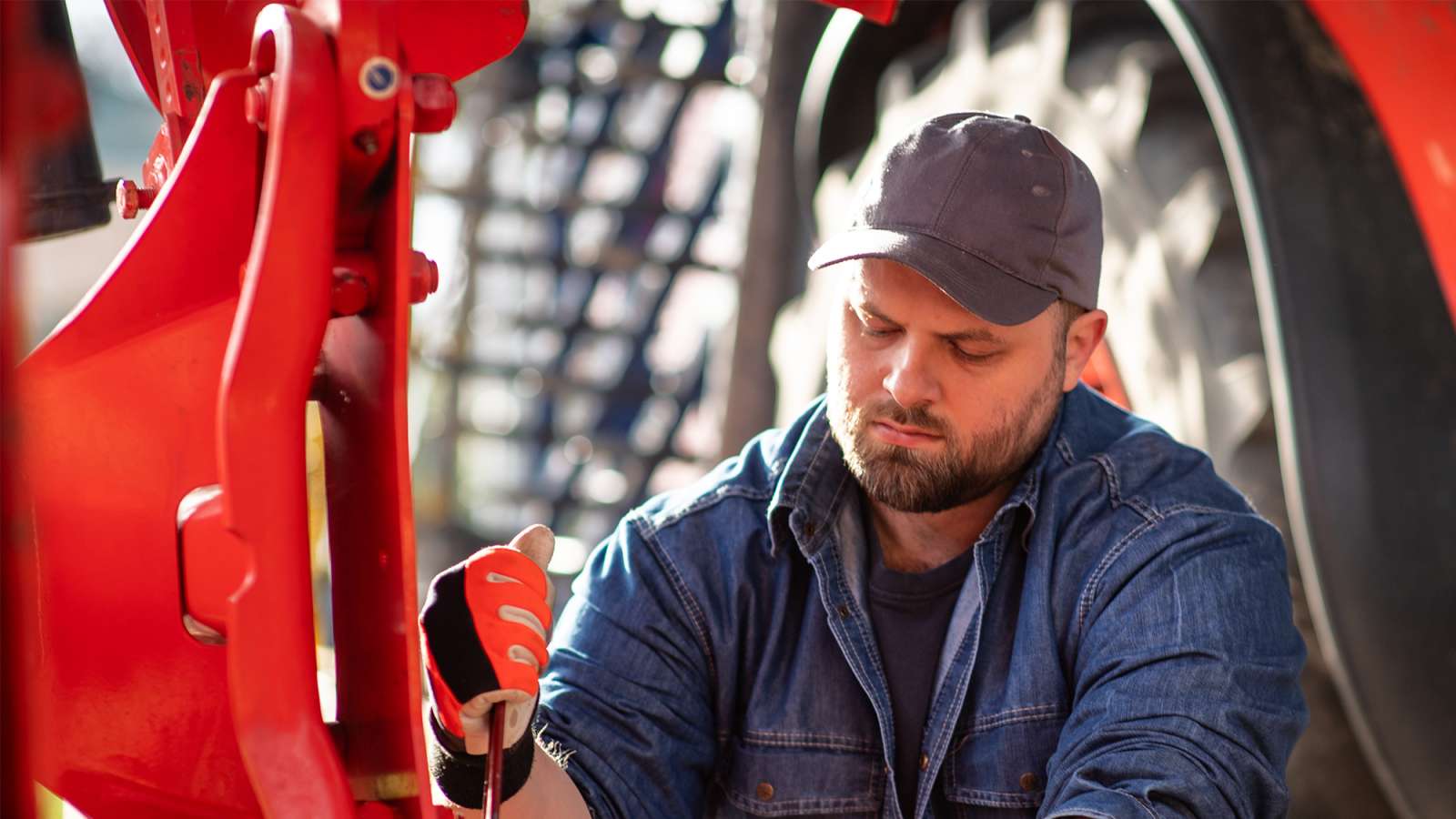 Bearded man in denim jacket and cap inspecting a red handheld diagnostic tool while kneeling beside a large red tractor tire.