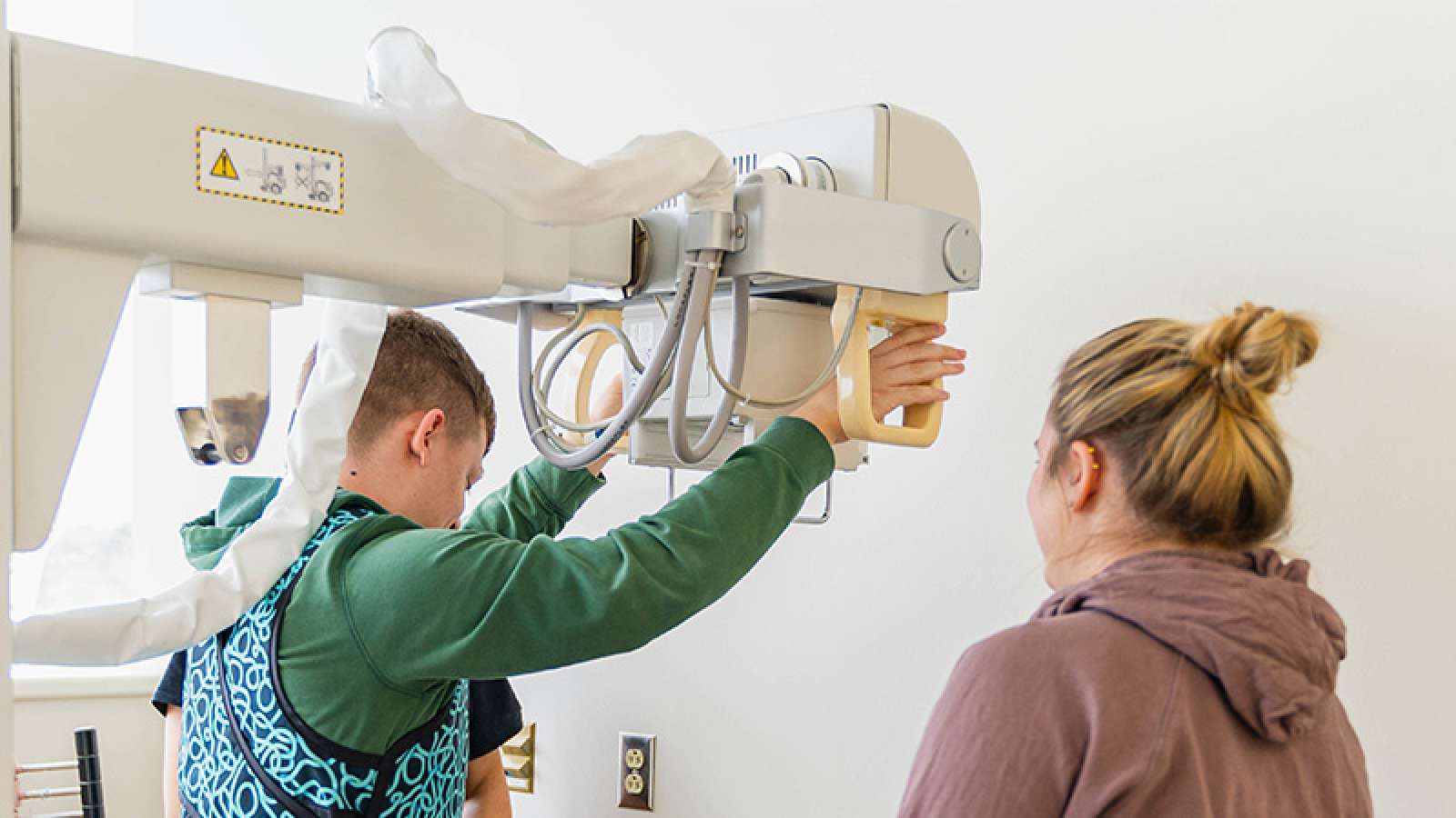 Radiology technician in lead apron adjusts overhead imaging device in a bright clinical room.