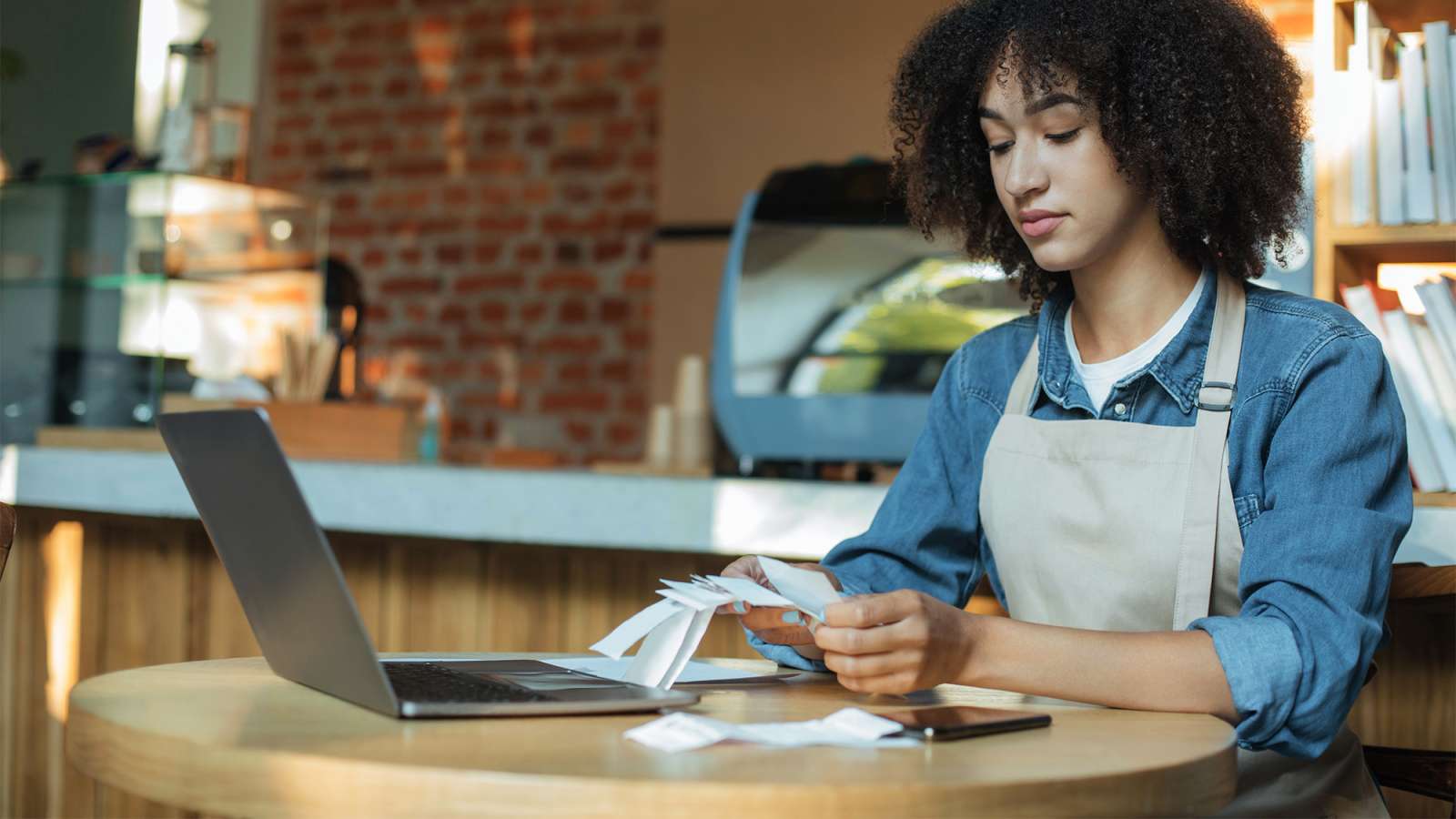 Focused young woman sitting at café counter, working on laptop with notebook, smartphone, and glasses.