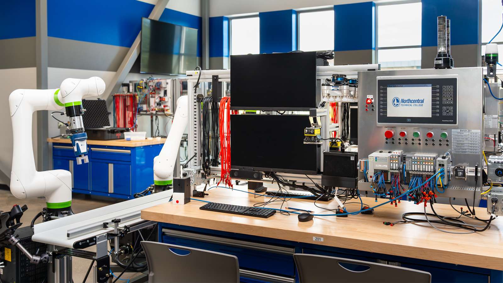 Electronics and robotics workbench with white articulated robotic arm, central monitor, circuit boards, and tools.