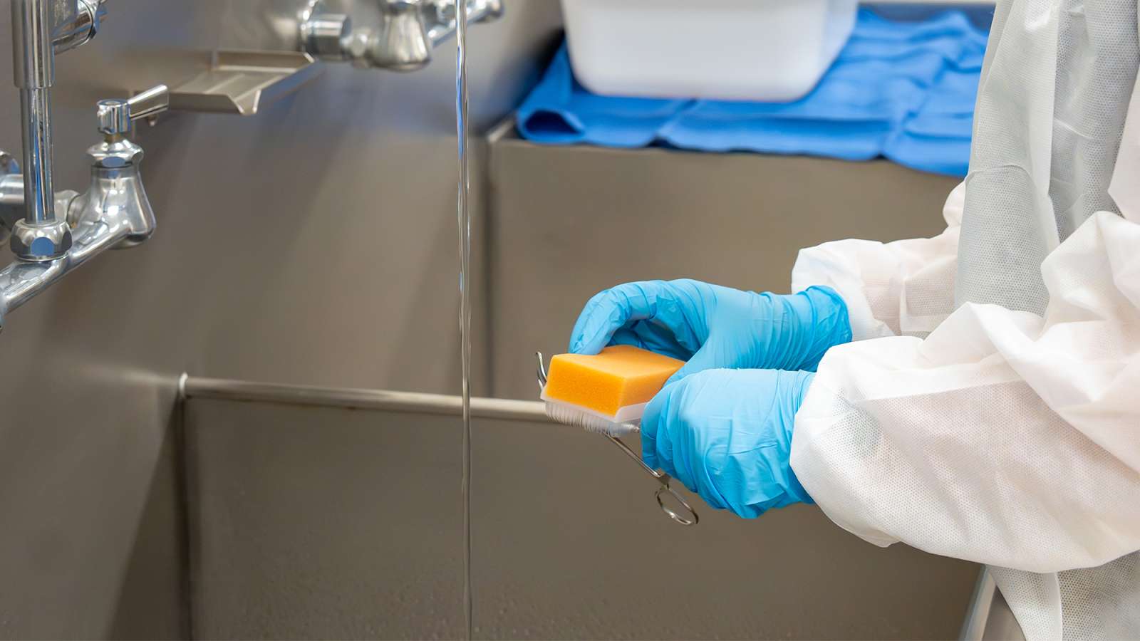 Person in white protective gown and blue gloves holds a small orange container under running water at a laboratory sink.