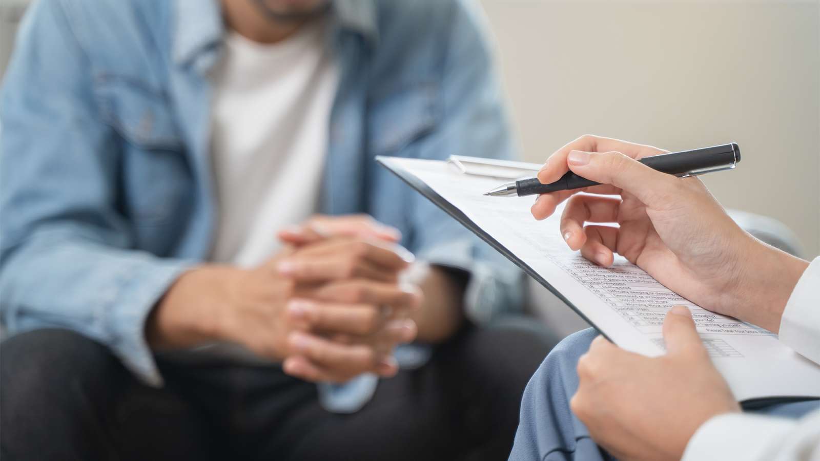 Clinician’s hand writing on clipboard while seated patient in blue denim jacket sits opposite in a counseling session.