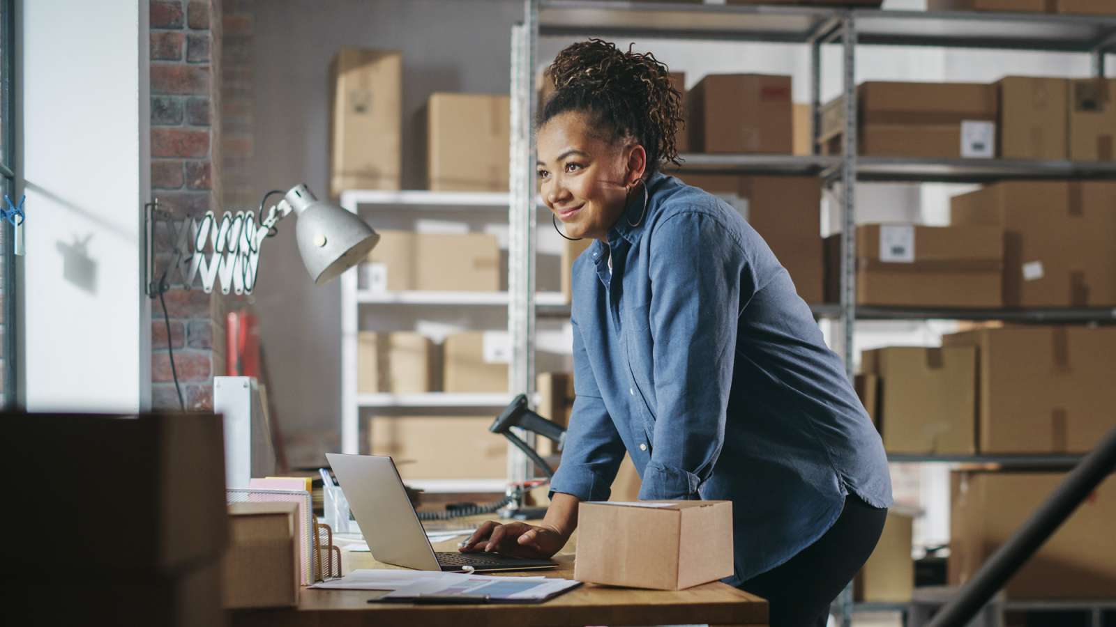 Smiling woman in denim shirt leans on packing table, typing laptop amid cardboard boxes and metal shelving in a warehouse.