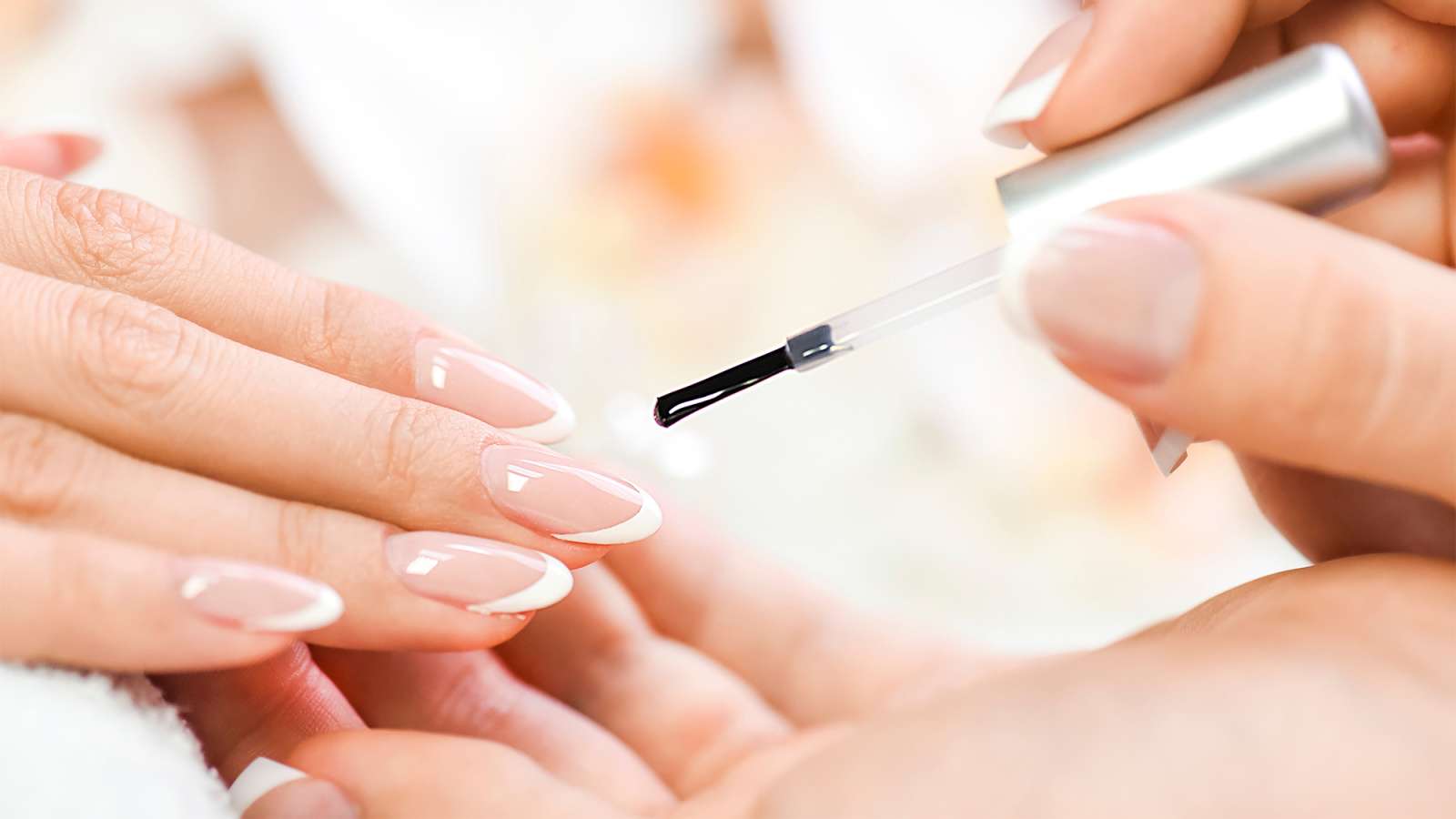 Hands receiving a French manicure: technician applies clear topcoat with a brush to neatly shaped pink nails with white tips.