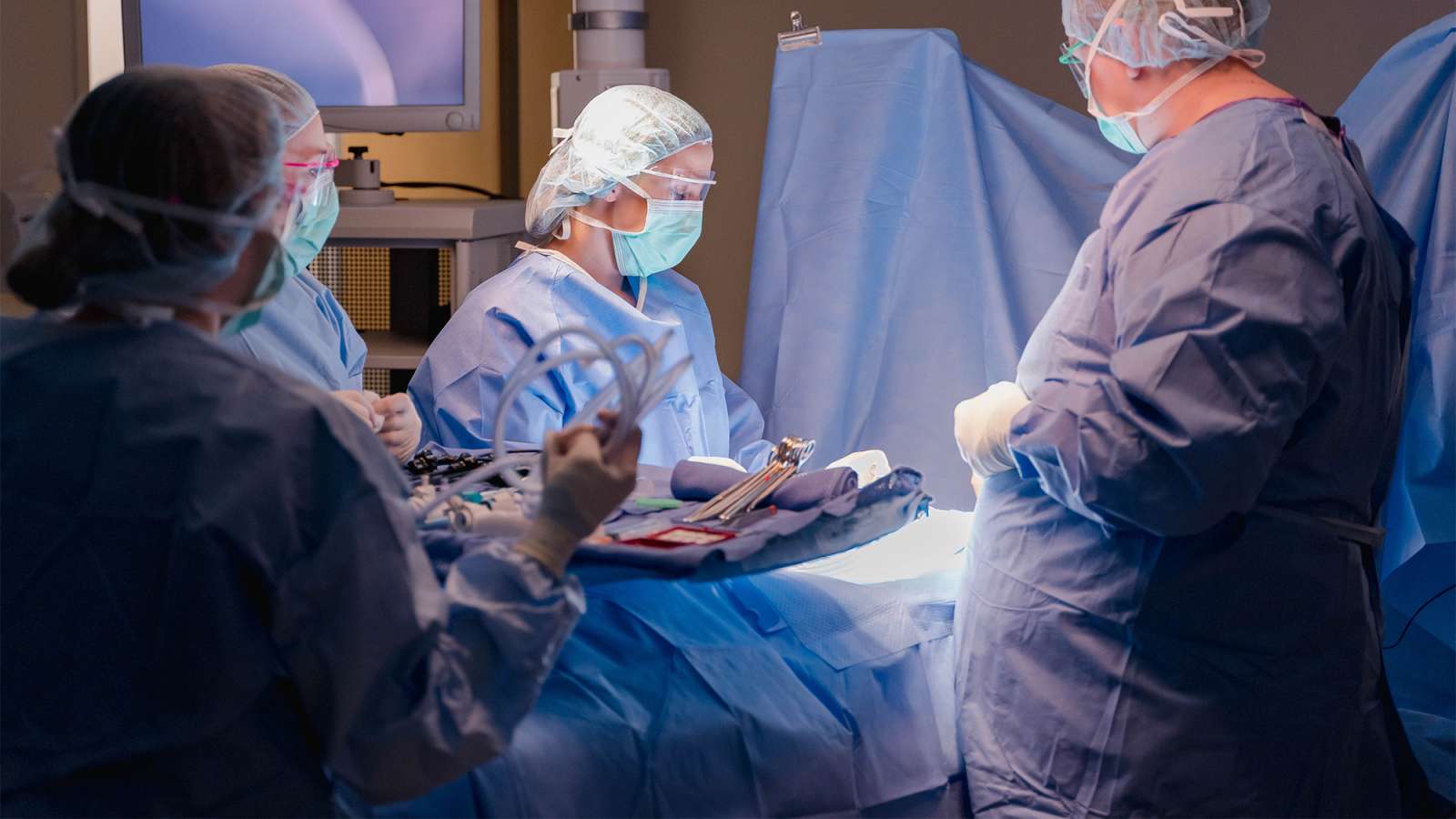 Surgical team in blue scrubs and masks performing an operating-room procedure under bright lights.