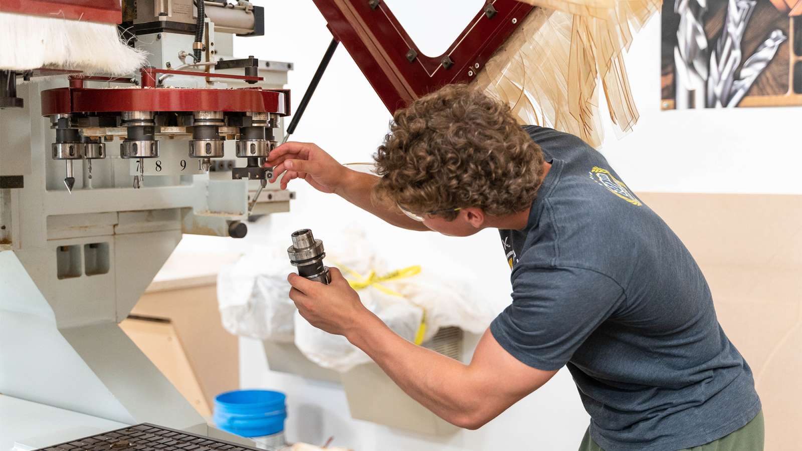 Man in dark shirt leans over a large industrial drill press, adjusting a metal component with tools in a cluttered workshop.