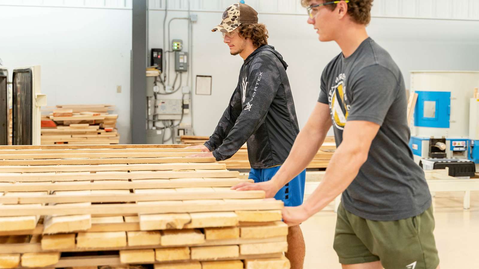 Two young men wearing safety glasses, one wearing blue gloves, moving a cart with a large stack of wood beams piled on it.