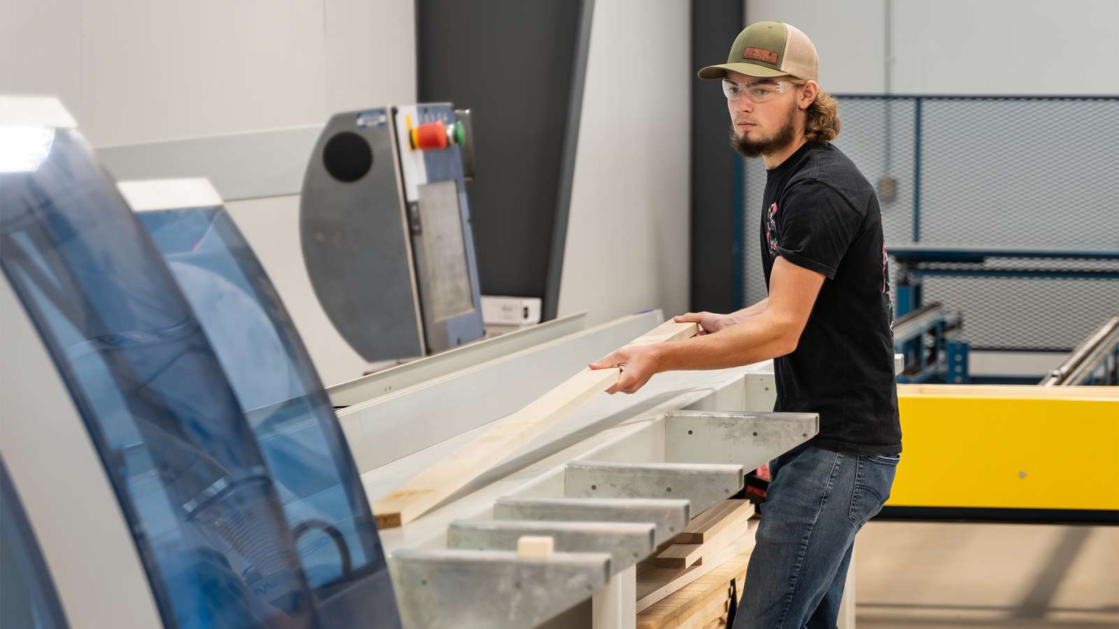 Young man wearing cap and headphones pushes long wooden board through an industrial cutting table in a bright workshop.