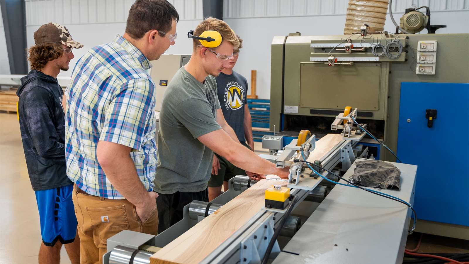 Three people in a workshop gathered around a long blue industrial machine; a man wearing yellow earmuffs adjusts material.