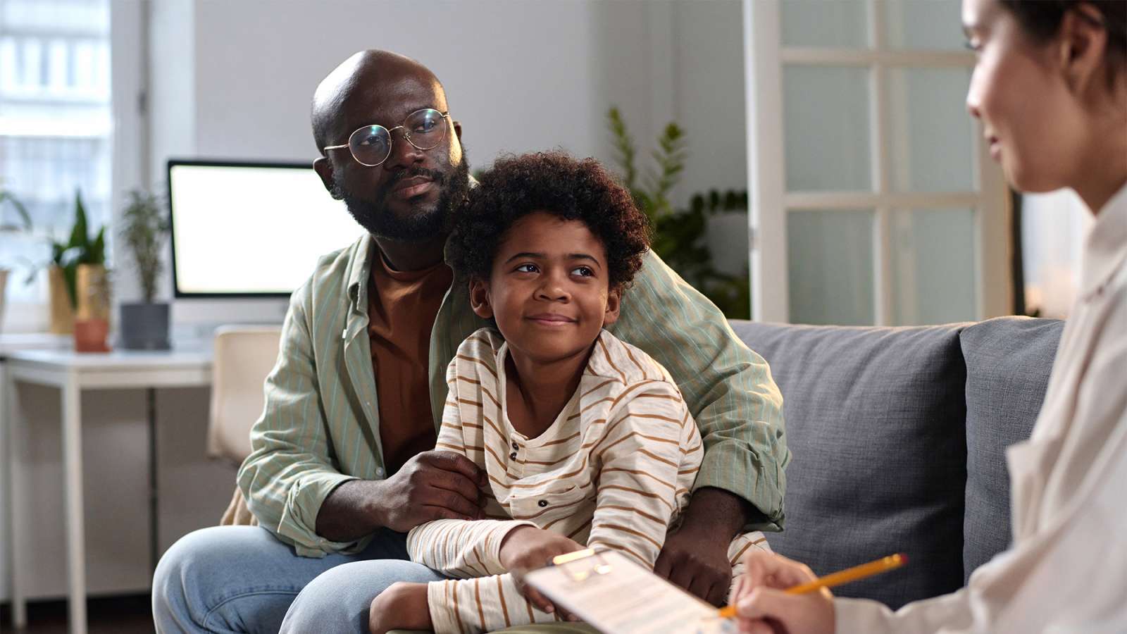Black father and young son sit on a living-room couch; father gently holds son as both engage with a female therapist.