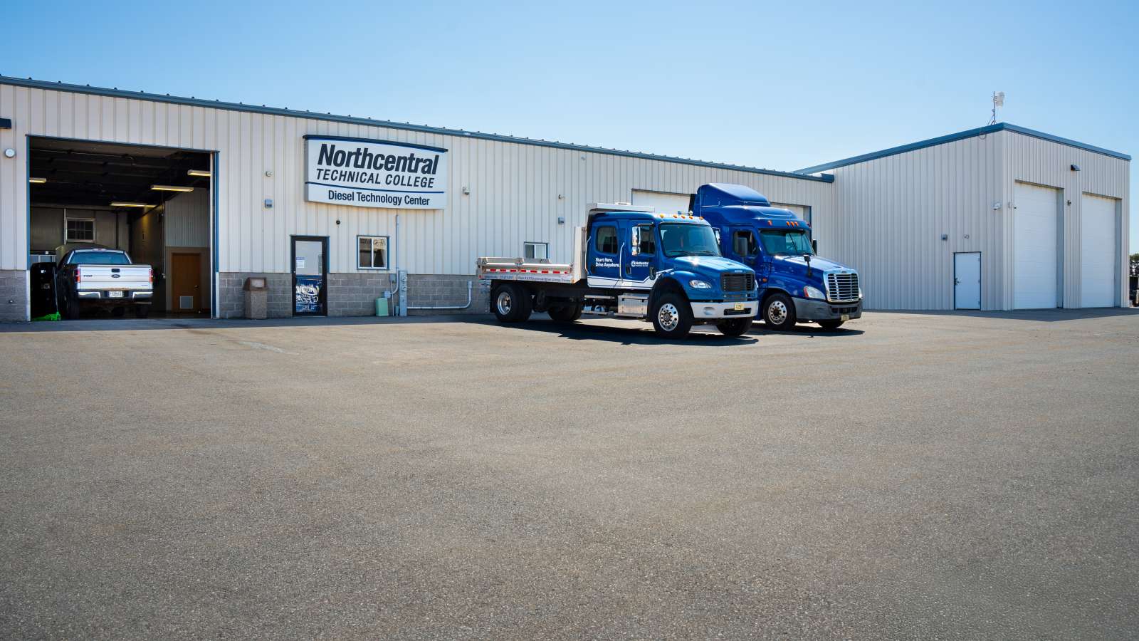 Two training trucks parked outside the front of the Diesel Technology Center with one bay open and two bays closed..