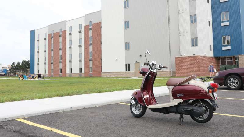 Exterior of Timberwolf Suites with students gathering, as seen from the parking lot. A moped is in the foreground. 