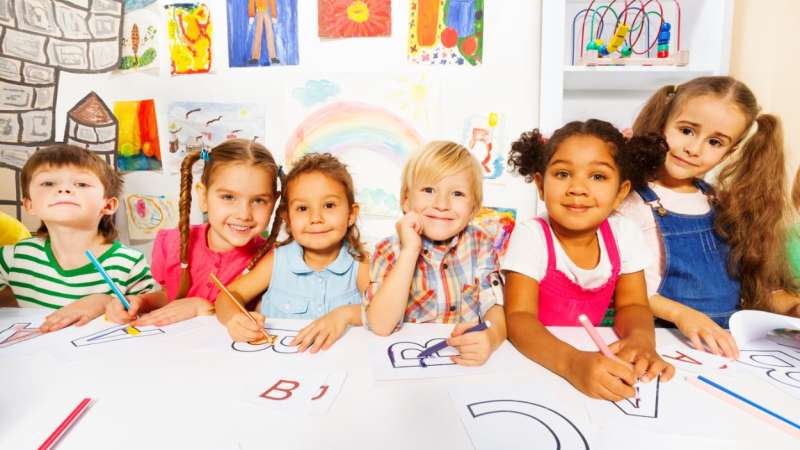 Young children in a classroom are sitting at a table drawing shapes on a large piece of art paper