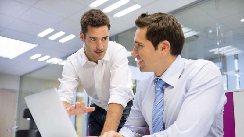 Two businessmen discuss something in a conference room while looking at a laptop screen.